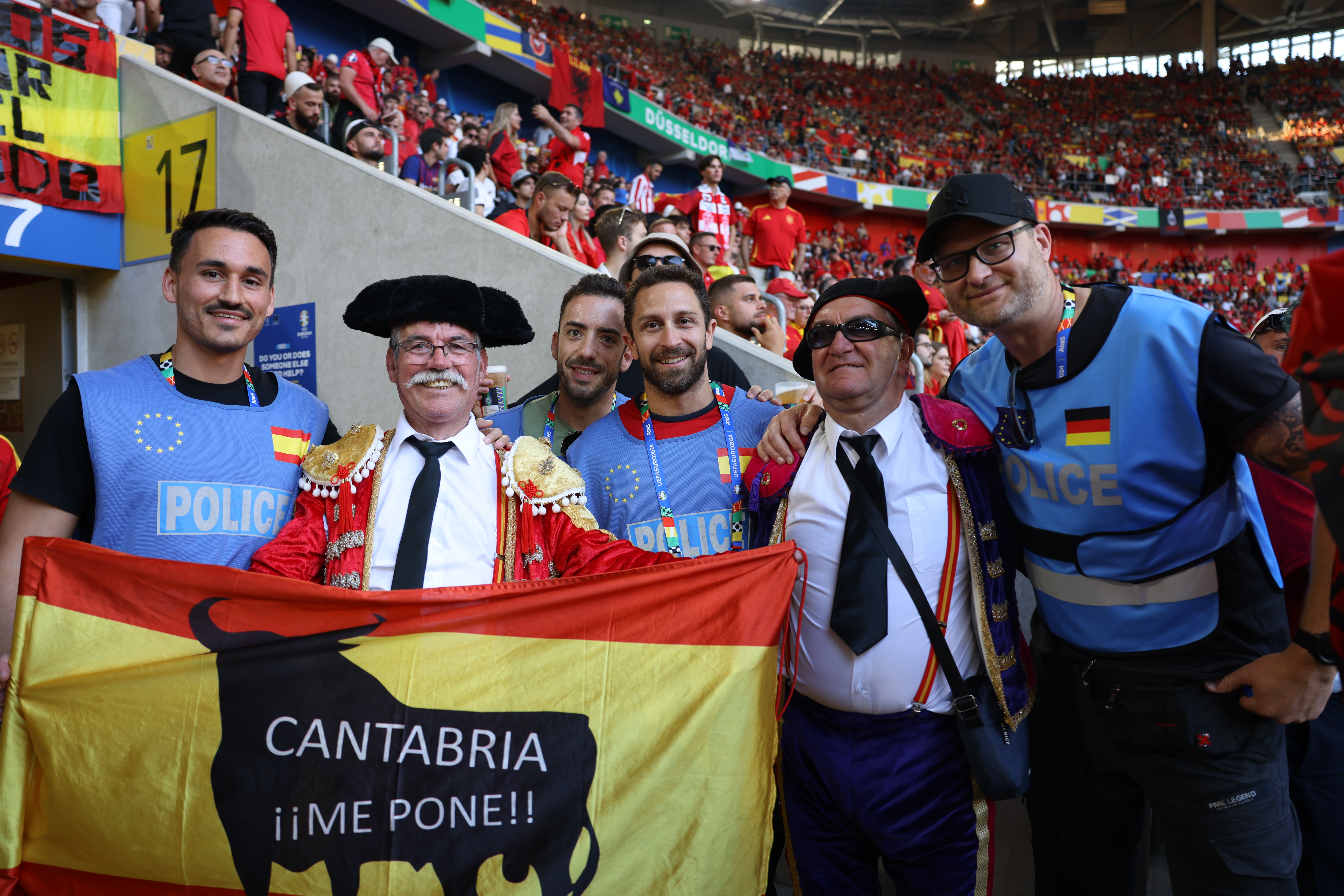 In einem vollen Fußballstadion stehen als Stierkämpfer verkleidete Fans und Polzisten aus Spanien und Deutschland zusmmen. 