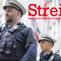 Zwei Polizisten stehen vor dem britischen und dem US-amerikanische Generalkonsulat in Düsseldorf. Sie bewachen es. Beide tragen Uniform, ihre Blicke gehen zum linken oberen Rand des Bildes. Im Hintergrund ist verschwommen das Gebäude und Fahnen zu sehen.