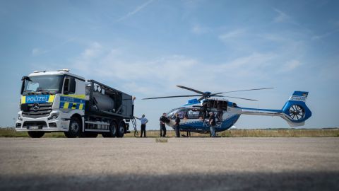 Auf dem Flughafengelände stehen der Tankwagen und ein Polizeihubschrauber nebeneinander. Davor stehen Mitglieder der Polizeifliegerstaffel. Im Hintergrund ist eine Wiese zu sehen und ein blauer Himmel.