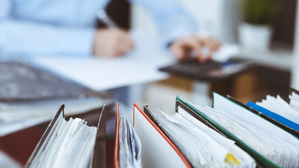 Aktenordner im Vordergrund, eine Person, die Papiere bearbeitet im Hindergrund